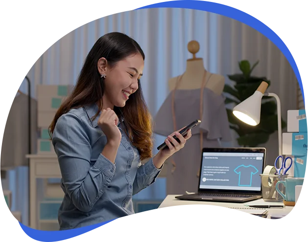 A woman seated at a desk is engaged with her phone, focused on the screen while surrounded by a tidy workspace.